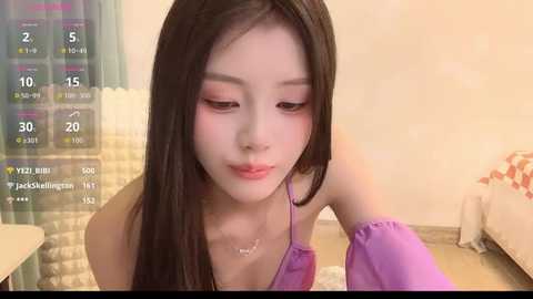 Video of an East Asian woman with long black hair, fair skin, and delicate features, wearing a purple top, in a bedroom with a digital clock displaying time.