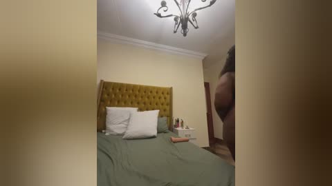 A video of a modern bedroom with a mustard-yellow tufted headboard, green bedspread, white pillows, and a beige wall. A chandelier hangs from the ceiling, and a person's lower body is partially visible in the corner.