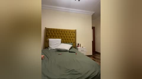 Video of a minimalist, beige-walled bedroom with a green bedspread, white pillows, a mustard-yellow upholstered headboard, a nightstand, and a wooden door.