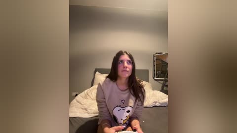 Media: Video of a young woman with long dark hair, wearing a gray sweater with a Snoopy design, sitting on a bed in a dimly lit room, holding a controller.