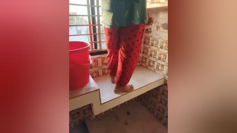 Media: Video of a person in red pants and green shirt, standing on a white ledge with a red bucket beside them, against a tiled wall with a window in a bright, possibly sunny room.