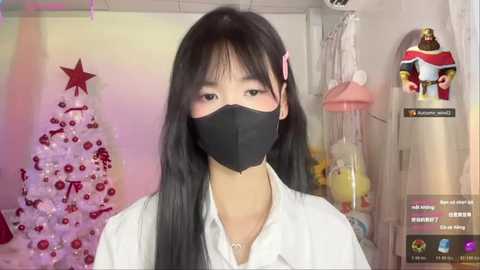 A video of an Asian woman with long black hair and a black mask, wearing a white shirt, standing in a festive room with a decorated Christmas tree and Santa figurines.