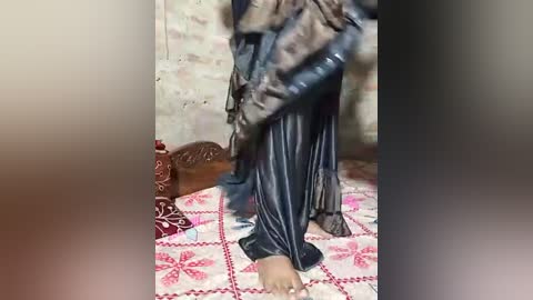 Media: Video of a person in a dark, flowing robe, standing on a patterned red and white rug in a room with a beige, textured wall and a wooden stool.