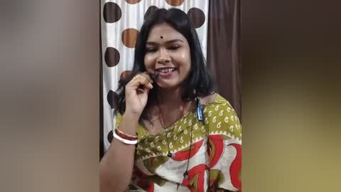 Video of a smiling South Asian woman with dark skin, medium-length black hair, and a bindi, wearing a mustard-yellow sari with red and white patterns, holding a phone to her ear.