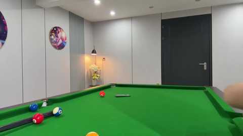 Media: Video of a modern, well-lit pool room with a green felt pool table, colorful balls, and a black door. Framed family photos adorn the white-paneled walls, and a lamp casts a warm glow.