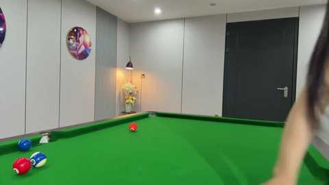 A video of a modern pool room with a green felted pool table, colorful balls, and white walls adorned with circular posters. A person is partially visible in the background.