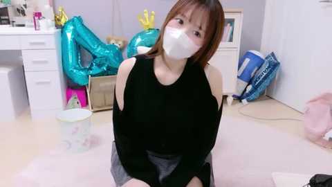Video of an Asian woman with shoulder-length brown hair, wearing a black top, white mask, and gray skirt, kneeling on a pink carpet in a cluttered room with balloons, a white dresser, and a vacuum.