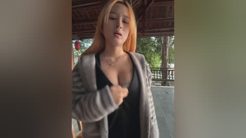 A video of an Asian woman with long, straight, blonde hair, wearing a black top and a gray cardigan, standing under a wooden pavilion, her eyes closed in apparent concentration.