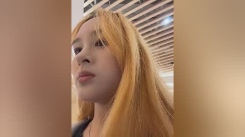 Video of a young Asian woman with long, straight, blonde hair, wearing a black top, standing in a modern, wooden-beamed room. She has a neutral expression.