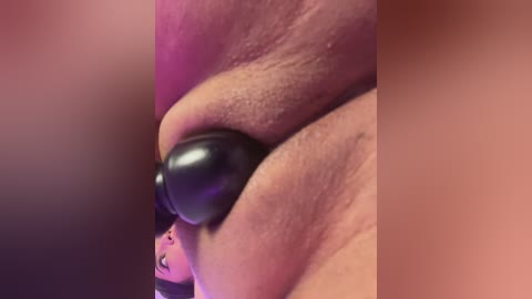 Close-up video of a person's left hand, showing a black bead inserted into the upper lip. The background is a gradient of purple and pink hues, creating a blurred, abstract effect.