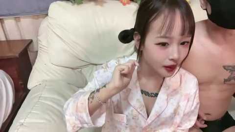 Video of a young Asian woman in a pink floral kimono, with red lipstick, sitting on a beige couch, holding a piece of paper. A shirtless Asian man stands behind her.