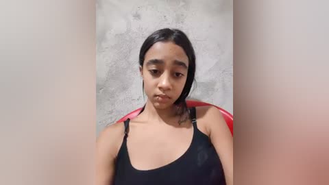 Video of a young woman with medium-brown skin, dark hair, and a serious expression, wearing a black tank top, sitting against a textured concrete wall with red chairs in the background.