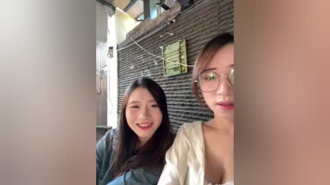 A video of two Asian women smiling, one with long dark hair, the other with short brown hair and glasses. They are indoors, in front of a brick wall with green signs.