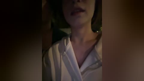 A dimly lit video of a young woman with fair skin, wearing a white blouse, partially obscured by shadowy figures in the background.