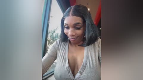 Video of a smiling Black woman with shoulder-length black hair, wearing a gray cardigan, standing in a modern, sunlit room with large windows and red curtains.