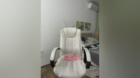 Video of a modern, white, reclining massage chair with pink flowers on the seat, in a minimalist, light-colored room with a bed and air conditioning unit in the background.