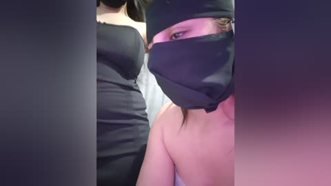 Video of two women, one wearing a black mask and black dress, the other with visible cleavage in a black strapless dress, partially visible in the background.