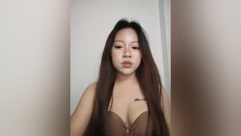 A video of a young Asian woman with long, straight black hair, wearing a strapless brown bra, standing against a plain white wall. Her expression is neutral.