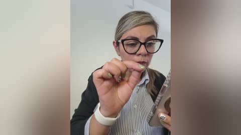 A video of a blonde woman with glasses, wearing a striped shirt and black jacket, taking a selfie in a well-lit room with blurred backgrounds.