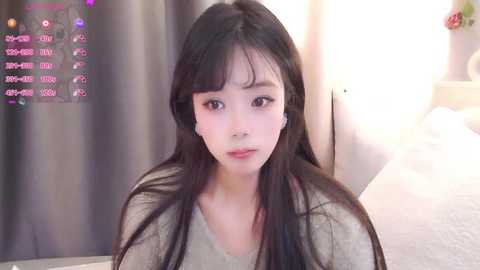 Video of a young Asian woman with long black hair, wearing a beige sweater, sitting indoors. Background includes a wall-mounted TV displaying a digital screen with text, and a blurred sofa.