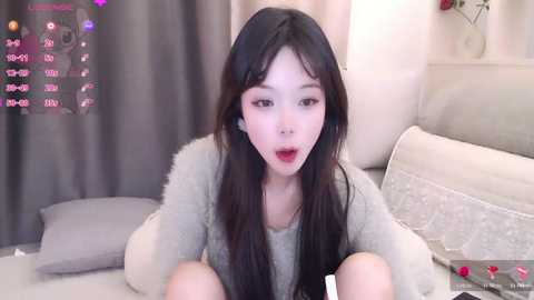 Video of an East Asian woman with long black hair, fair skin, and red lipstick, wearing a gray fuzzy sweater, sitting on a bed. The background features gray curtains, a white pillow, and a lamp.
