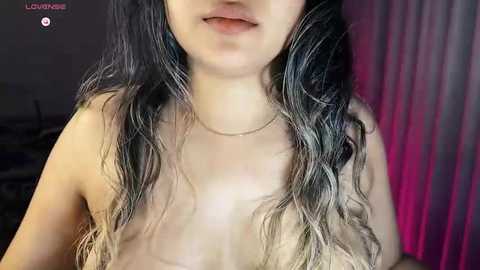 Video of a young woman with long, wavy, dark hair, pale skin, and small breasts, wearing a thin necklace, taken indoors with dim lighting.