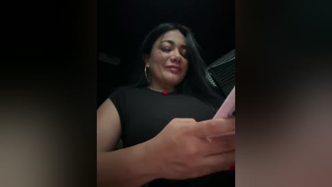 Video of a Latina woman with long black hair, wearing a black top and hoop earrings, holding a pink phone, smiling, in a dimly-lit room.