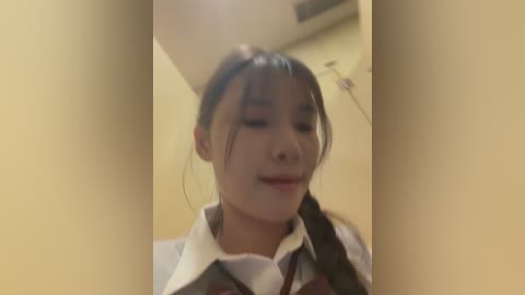 Video of an Asian woman with long black hair tied in a ponytail, wearing a white collared shirt with a brown vest, smiling. Background shows a beige wall and ceiling.