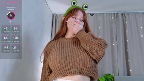 A video of a red-haired woman wearing a green frog hat and a knit brown crop top, covering her mouth with her hand. She stands in a modern room with gray curtains.