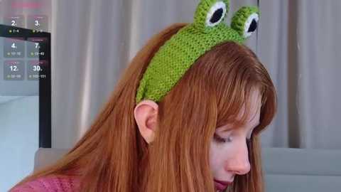 Video of a young woman with long red hair, wearing a green crocheted frog hat with large eyes, standing indoors with beige curtains in the background.