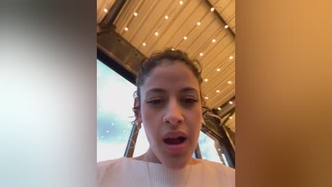 Video of a young woman with light skin and curly hair, wearing a white sweater, looking surprised, under a wooden, lit ceiling with visible beams.