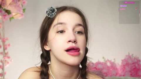 Video of a young woman with fair skin, brown hair in twin braids, wearing a grey hair clip and bold red lipstick. She's indoors with a pink floral backdrop, looking slightly to the left.