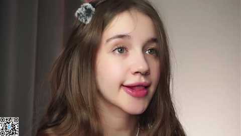 Video of a young Caucasian girl with long brown hair, wearing a silver hair clip, smiling. Background is blurred.