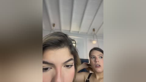 Video of two women in a modern room; one with long, dark hair, and the other with short, brown hair. The background features white ceiling beams and a blurred, neutral-colored wall.