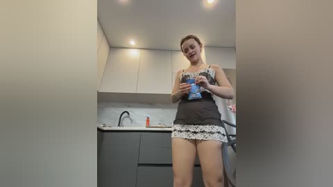 A video of a light-skinned woman in a black and white maid outfit, holding a blue bottle, in a modern kitchen with white cabinets and grey countertops.