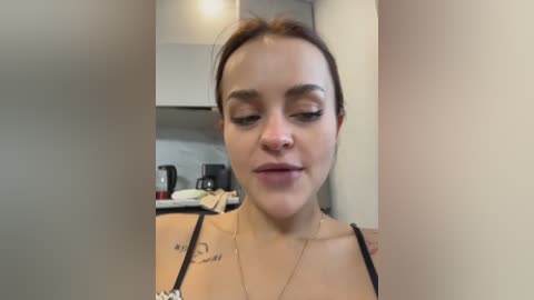 Media: Video of a light-skinned woman with brown hair, wearing a black top, standing in a modern kitchen with stainless steel appliances and white walls.