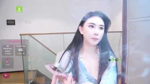 Video of a young Asian woman with long black hair, wearing a light blue blouse, standing in front of a glass railing, looking up.