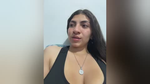 A video of a Latina woman with medium skin tone, long black hair, and large breasts, wearing a black halter top. She has a neutral expression and is standing against a plain, light-colored wall.