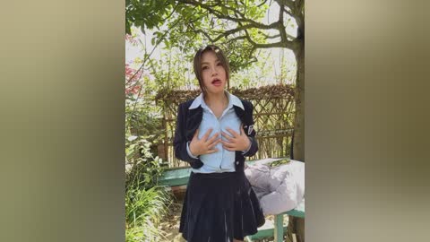 A video of a woman in a black school uniform, hands on her chest, standing outdoors in a garden with greenery and a white fence in the background.
