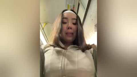 A video of a young woman with shoulder-length brown hair, wearing a white headband and a cream-colored jacket, smiling widely in a dimly lit bathroom with a yellow towel and a mirror in the background.