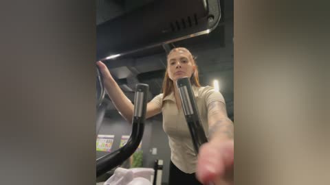 Video of a fit, fair-skinned woman with straight brown hair, wearing a beige shirt and black pants, working out on an elliptical machine in a modern gym with dark walls and exposed ceiling beams.