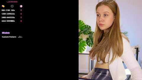 Video of a young Caucasian woman with long, straight, light brown hair, wearing a white top and black leather skirt, seated indoors with green plants in the background.
