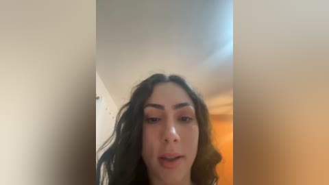 A video of a young woman with long, wavy black hair and fair skin, wearing subtle makeup. She has a neutral expression and is seen from the waist up. The background is blurred, showing a beige wall and a warm light source.