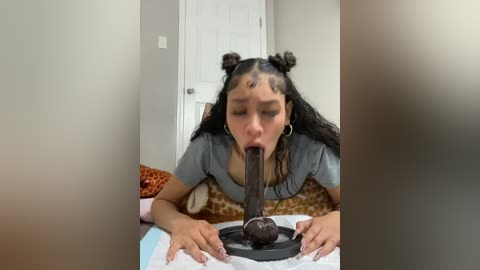 Video of a young woman with medium brown skin, wearing a grey top, kneeling on a bed, sucking a large, dark, dildo. Background shows a closed door and a patterned blanket.