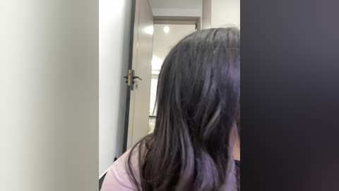 Video of a person with long, dark hair partially obscuring their face, standing in a modern hallway with beige walls and a partially open door.