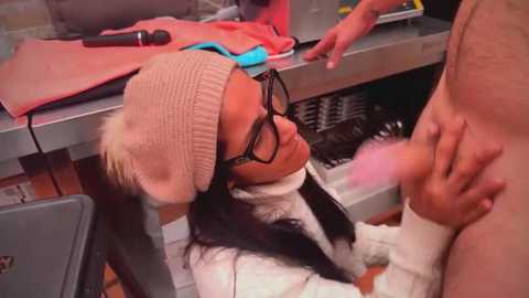 Video: Asian woman in glasses and knit hat, white sweater, kneeling, performing oral sex on a nude man in a dimly-lit, industrial kitchen with exposed pipes and a red towel.