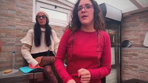 Video of two women in a tiled bathroom; one seated, wearing a beige sweater, black skirt, and glasses; the other standing, with curly hair, wearing a red sweater, black stockings, and glasses.