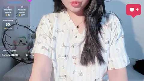 Video of an Asian woman with long black hair, wearing a white T-shirt with cartoon car print. Background features a plush toy and a red heart emoji.
