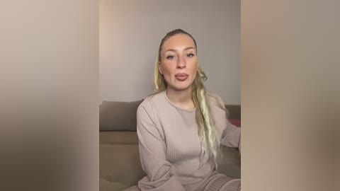 A video of a blonde woman with fair skin, wearing a beige knit sweater, sitting on a beige couch against a plain background.