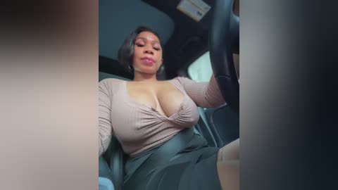 A video of a curvy, dark-skinned woman with large breasts wearing a low-cut, beige top and a grey skirt, sitting in a car, holding the steering wheel.